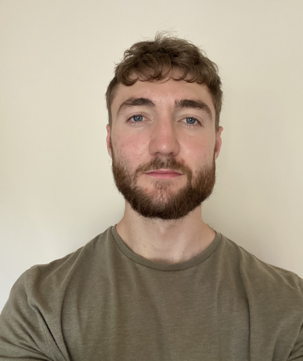A portrait photograph of a man with short brown hair, trimmed beard, and blue eyes, wearing an olive-green t-shirt, standing against a plain light-colored background